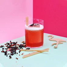 Carica l&#39;immagine nel visualizzatore di Gallery, Cocktail served with bagasse straws on a colorful background with scattered herbs and a wooden flamingo decoration.